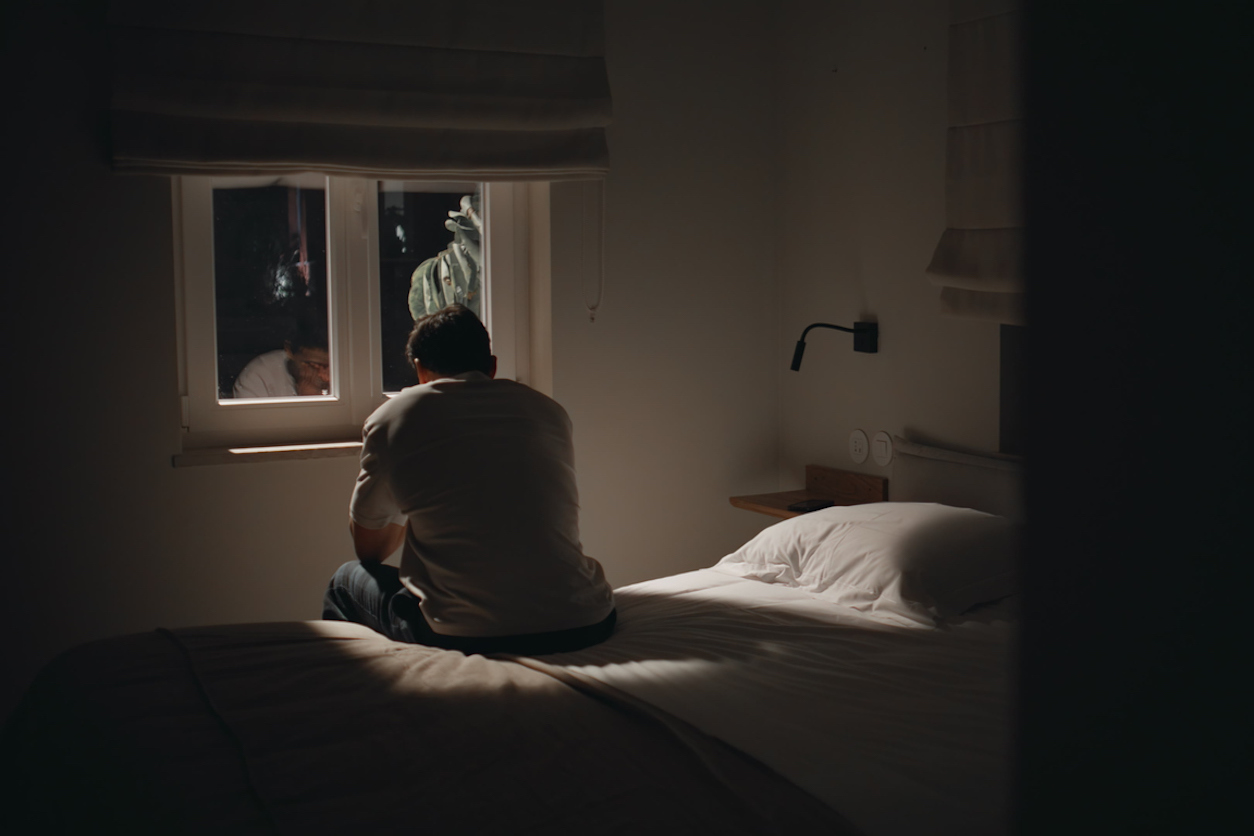 A man sitting on his bed, facing away from the door, at home in a dim-lit room looking very lonely.